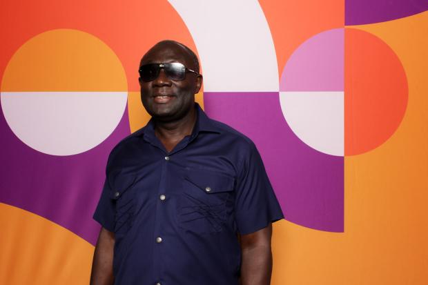 Kobena Bonney stands in front of a colorful backdrop of geometric shapes. He's wearing dark sunglasses and a dark blue button-down shirt.