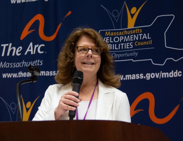 Mary McGeown, a white woman with light brown hair wearing a white shirt and blazer and glasses stands at a podium speaking.