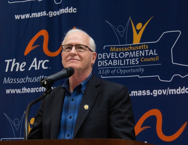 Michael Barrett, a caucasian man with short white hair wearing a blue and black shirt and black blazer stands at a podium speaking.