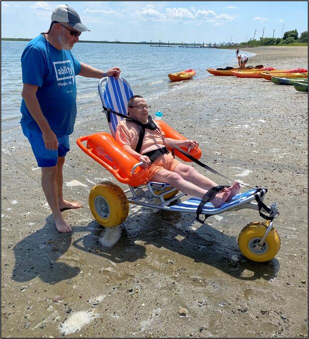 A  person is sitting in a floating beach wheelchair after floating in the water. A person is pushing them from behind.
