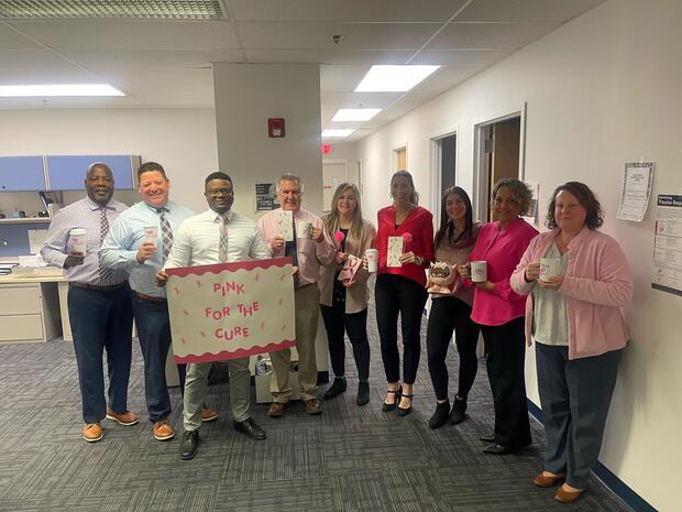 A group of eight people stands in an office, holding pink-themed items, with a sign reading "PINK FOR THE CURE."