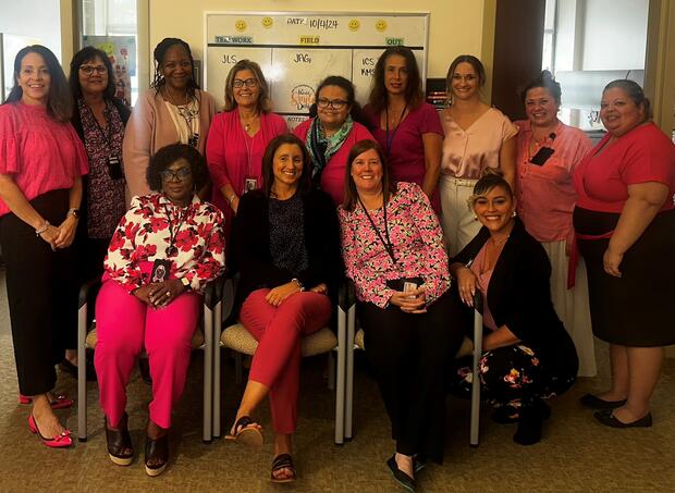 A group of fifteen women in pink and black clothing, with some seated and others standing, in front of a whiteboard.
