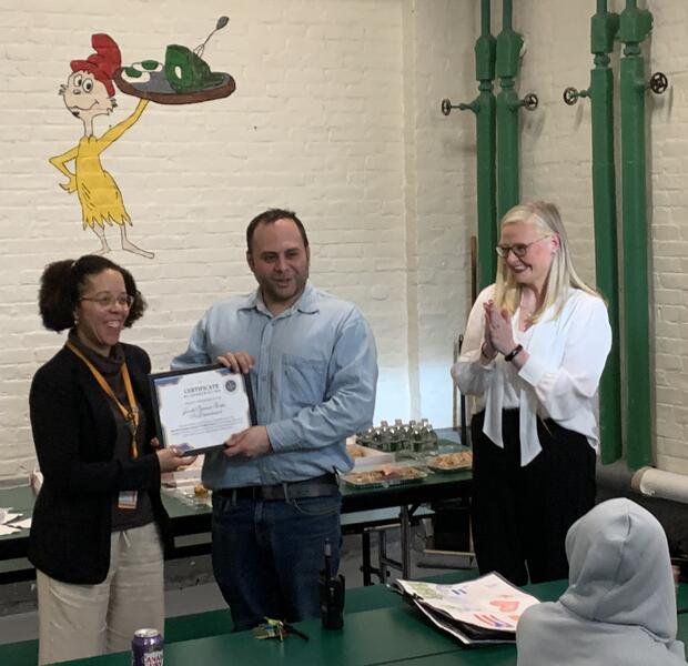 Three people in a room, one receiving a certificate, others clapping and smiling.