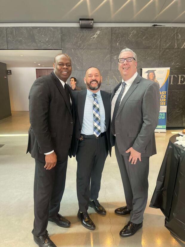 Three men in formal suits smiling and standing together indoors.