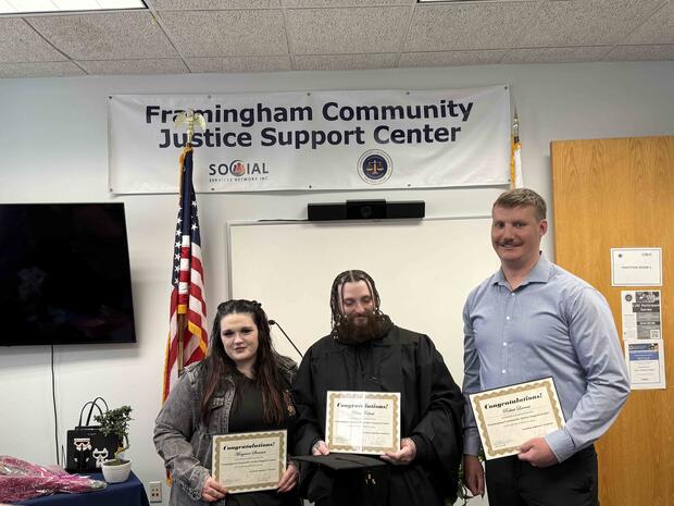 Three individuals holding certificates in front of a "Framingham Community Justice Support Center" banner.