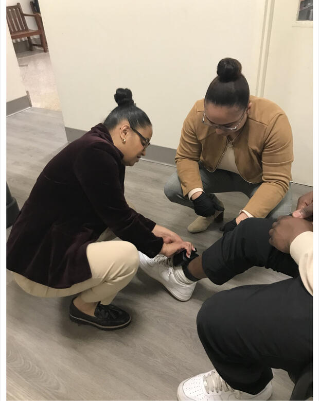 Two people adjusting an ankle device on a seated person's leg in a hallway.