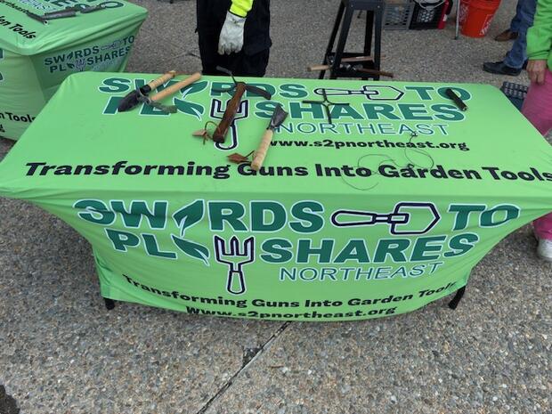 A table with a green cloth featuring "Swords to Plowshares Northeast," transforming guns into gardening tools theme, with tools displayed on top.