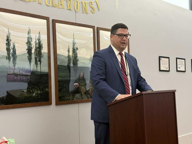 Man in a suit speaking at a podium with landscape paintings and framed texts in the background.