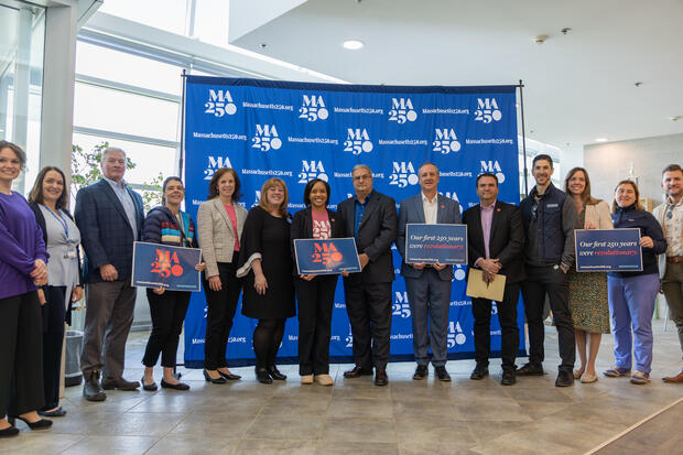 Attendees at kickoff event take a group picture standing in front of the Massachusetts 250 step and repeat banner. 