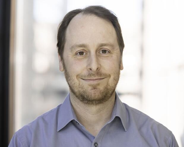 Matt with a beard smiling in front of a window and wearing blue top