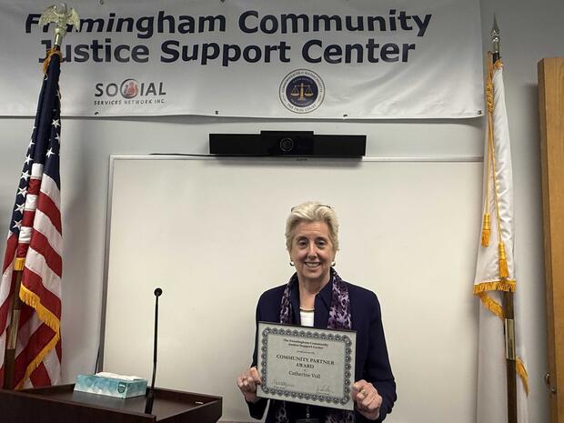A person holds a certificate in front of a "Framingham Community Justice Support Center" banner, between two flags.