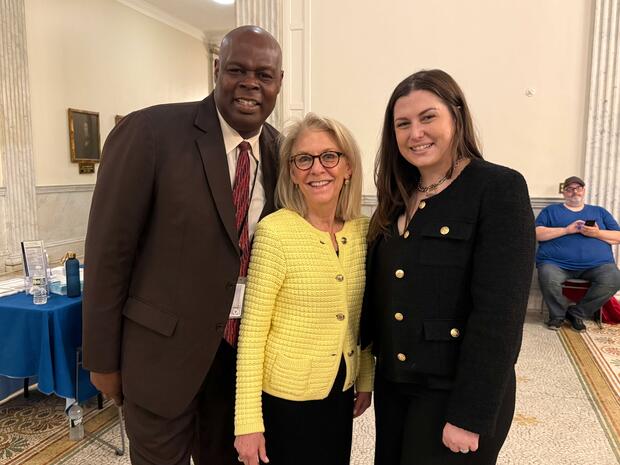 Three people smiling in a formal setting with a seated person in the background.
