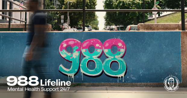 988 written on wall in graffiti with person walking by on the sidewalk. Text: 988 Lifeline, mental health support 24/7