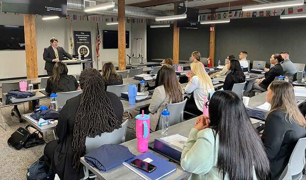 A presenter speaks to a classroom of students seated at tables.