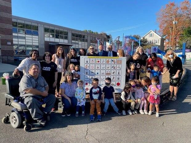 Group of children and adults gathered outside around a large communication board containing several icons. Playground and school building in background.