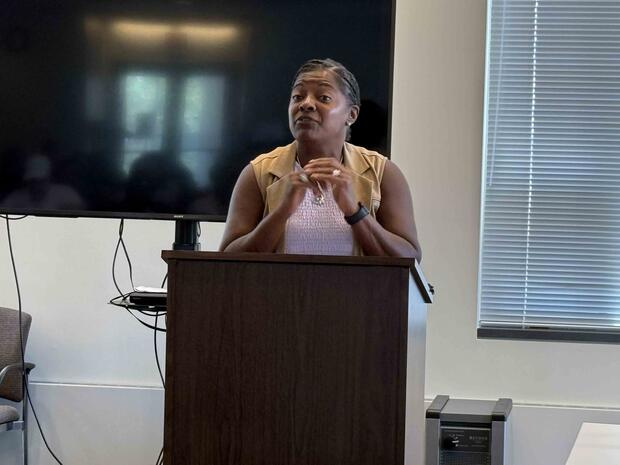 Person speaking at a lectern with a TV and window blinds in the background.