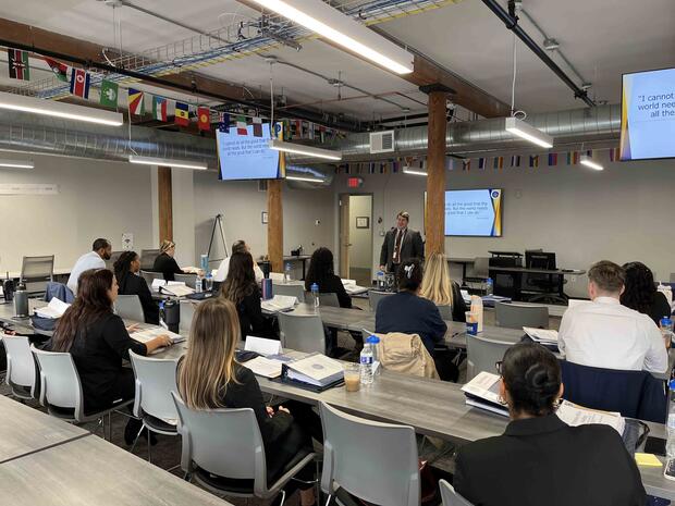 A classroom with people seated facing a presenter, flags overhead, and slides visible on monitors.