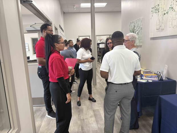 A group of people in an indoor office setting engaged in conversation, with brochures on a nearby table.