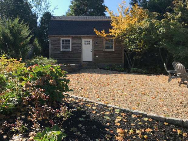 A pea gravel patio next to a small cabin and surrounded by trees, shrubs, and grasses.