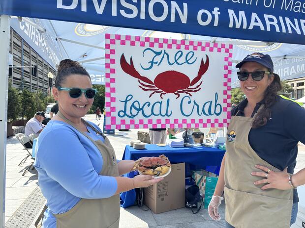 Two women hold a cooked crab next to a sign that reads "Free Local Crab". 
