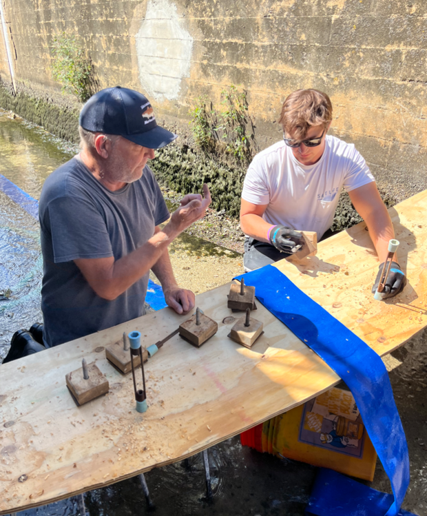 Two men stand in a fishway work on constructing parts on a large wooden plank. 