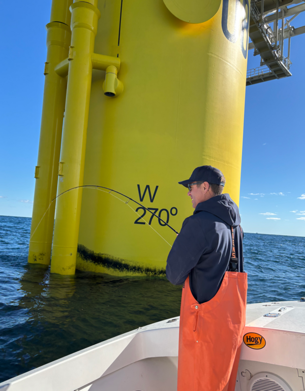 Man wearing orange Grundens fishes on a boat directly next to an offshore wind turbine. 