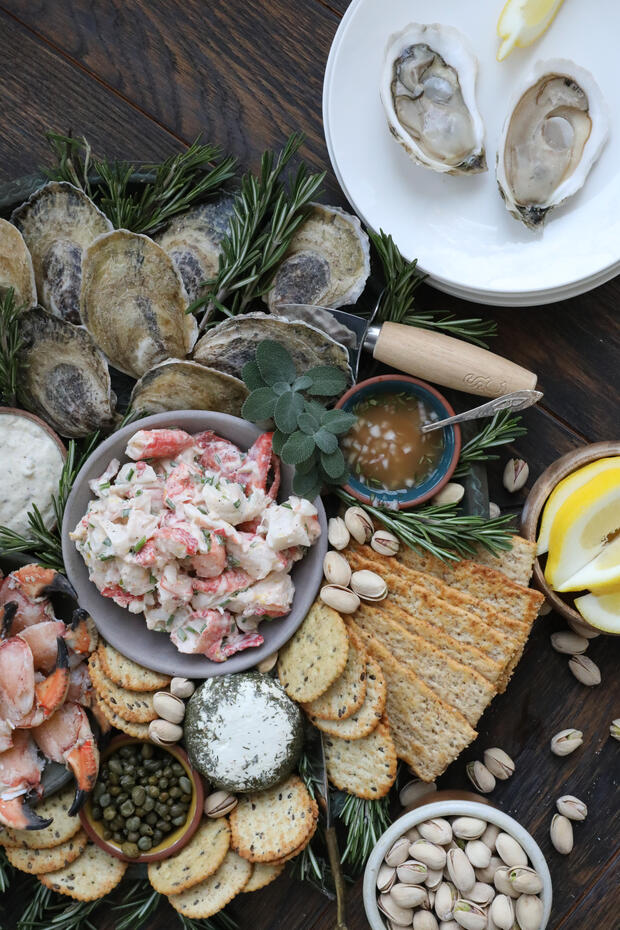 Serving platter with bowl of lobster salad, fresh oysters, and crackers and cheese on the side with sprinkled herbs throughout. 