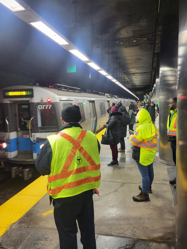 A train station platform filled with people, two MBTA employees in yellow safety vests watch the train and the platform.