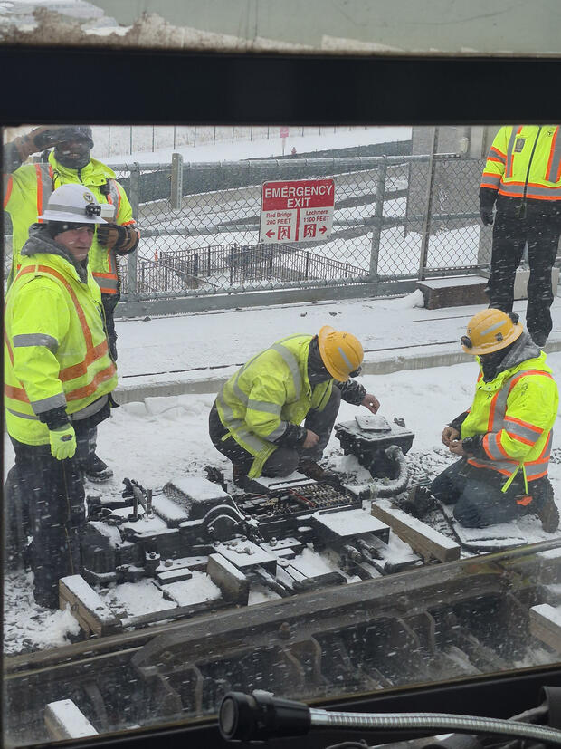 MBTA employees in yellow safety vests and hard hats fix a rail switch in snowy weather.