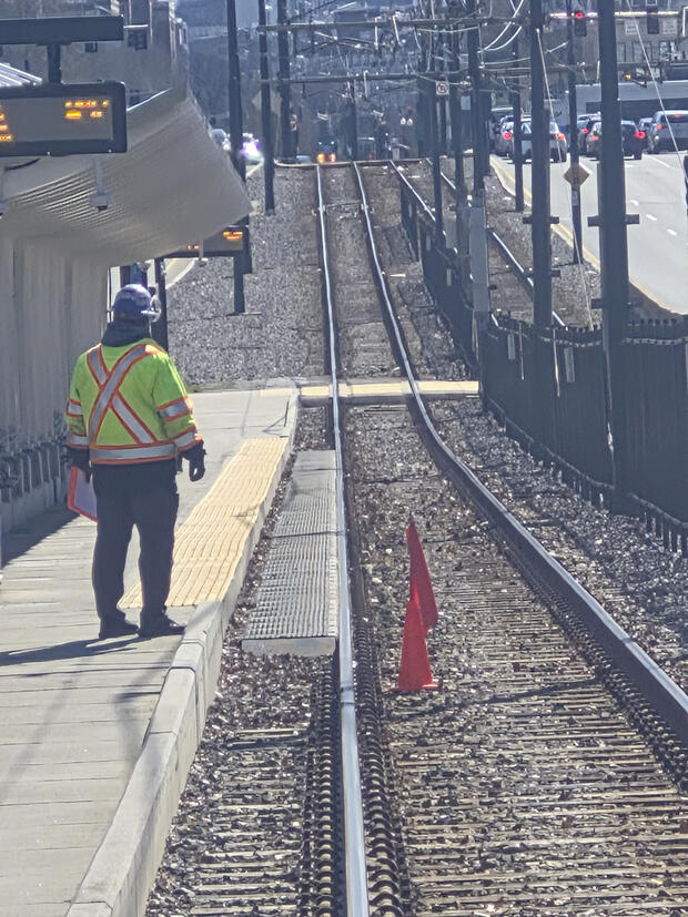 MBTA worker in a yellow safety vest stands on the rail platform watching the track area.