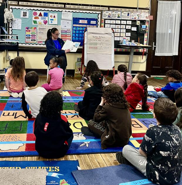 A person reading a book to children as they sit on a colorful mat decorated with letters of the alphabet.