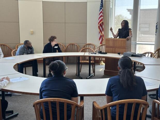 Students sitting at a circular table listening to a woman speaking at a podium.