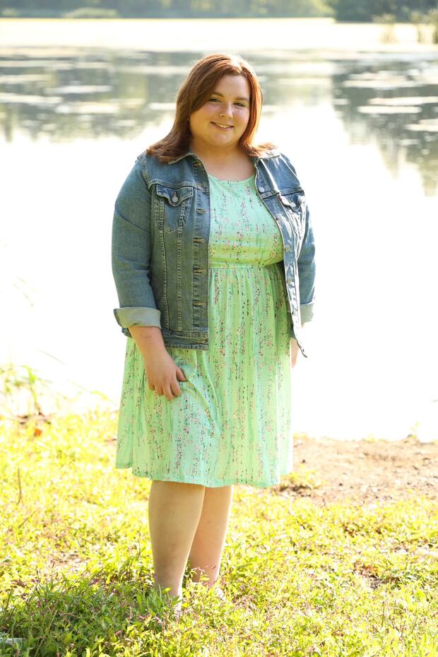 A person wearing a dress and denim jacket standing in grass in front of a body of water.