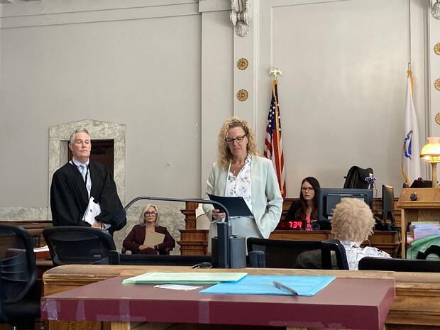 A courtroom scene with a judge and a woman speaking, accompanied by flags and digital clock.