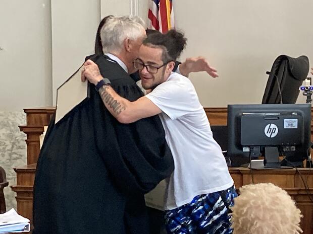 A man in casual clothes hugs a judge in a courtroom, with a computer and American flag visible in the background.