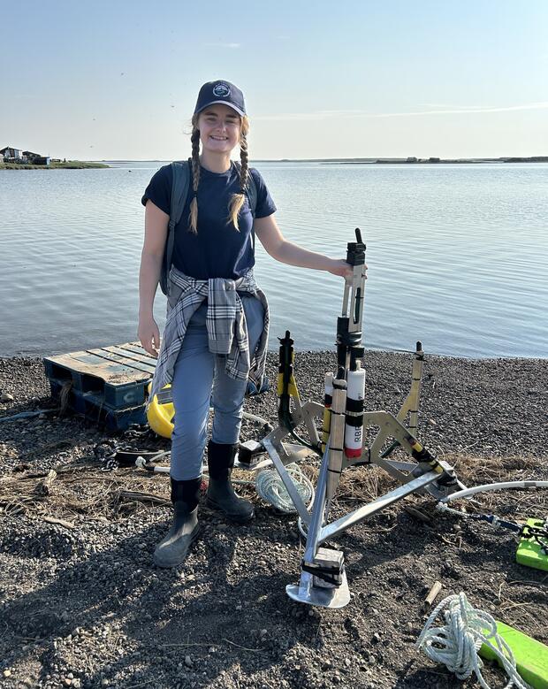 Woman stands at waterfront next to scientific instrument. 