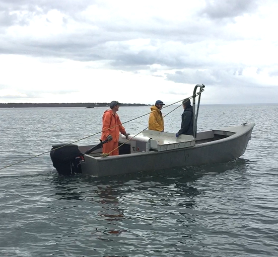 Three people stand on a small vessel, with lines appearing to tow something behind the boat below water. 