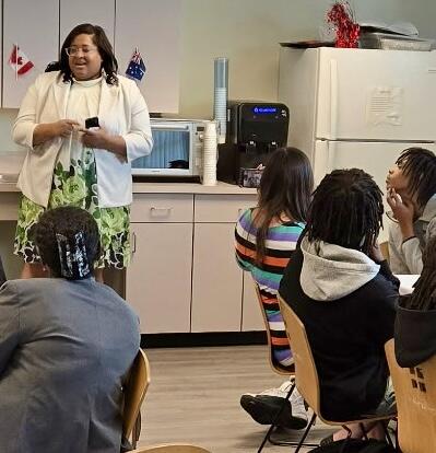 A woman speaking to a group of people seated in a cafeteria with a fridge and microwave in the background.