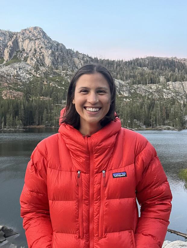 Woman wearing orange jacket smiles in front of mountain. 