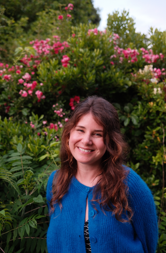 Woman in blue sweater smiles next to flowers. 