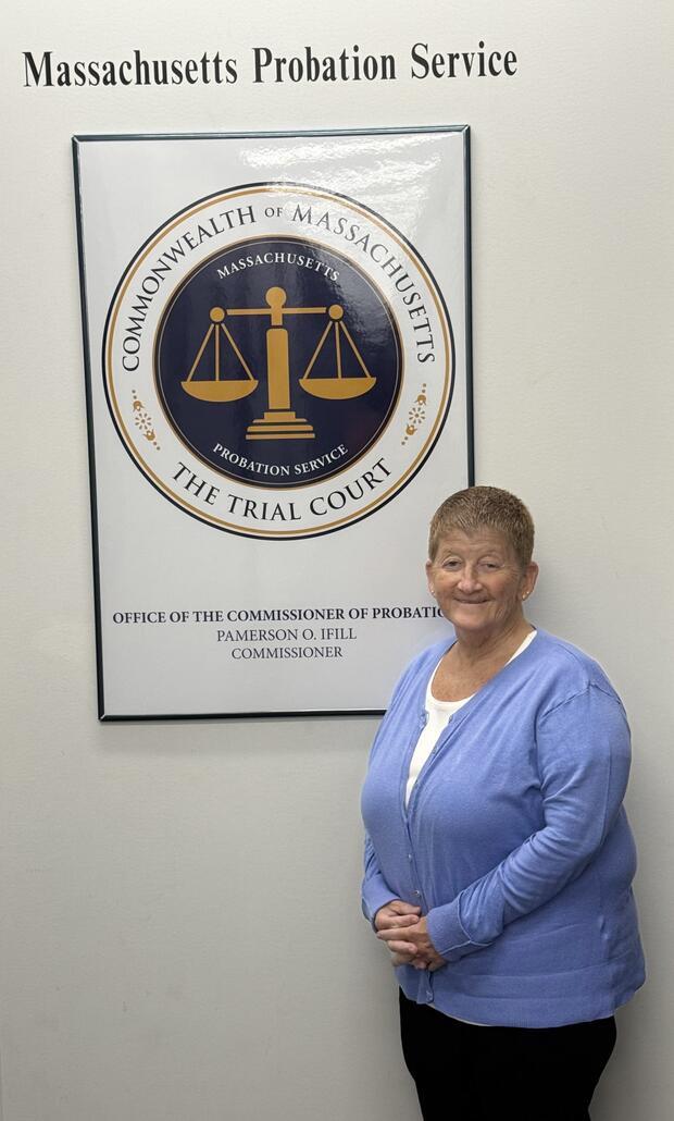 A woman stands beside a framed emblem of the Commonwealth of Massachusetts, which features scales symbolizing justice.