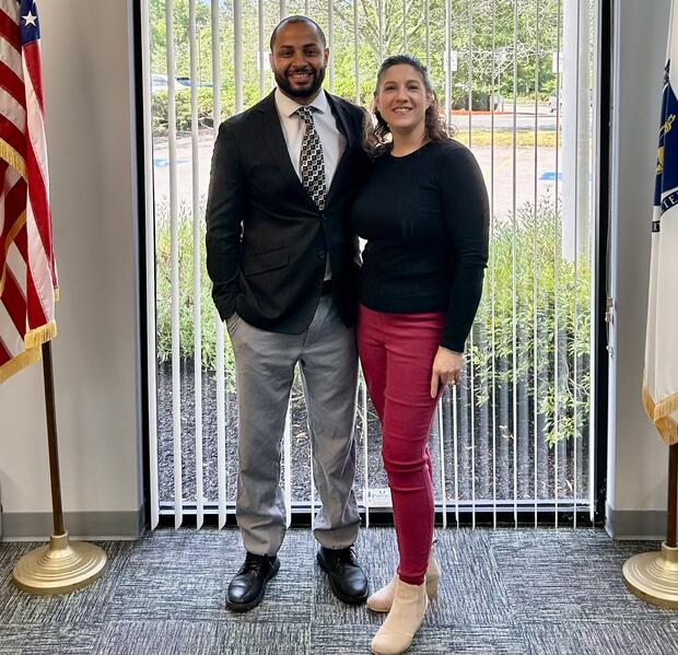 Two people standing in an office decorated with an American flag and the MA state flag.