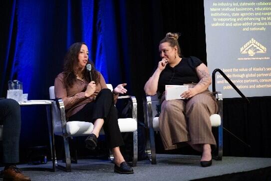 Two women sit on stage speaking into microphones. 