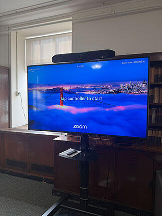 A wheeled monitor with a video-conferencing camera displays a Zoom start screen featuring a nighttime suspension bridge and city skyline.