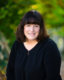 Smiling woman with shoulder-length dark hair wearing a black top against a green, leafy background.