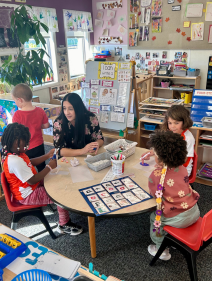 A teacher helps young children with arts and crafts around a table in a colorful classroom.