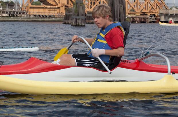 A photo of a young man sitting in a canoe with a large outrigger. He is holding a curved paddle in front of him with both hands.
