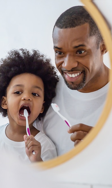A little kid brushing their teeth and their dad holding a toothbrush looking proud and happy