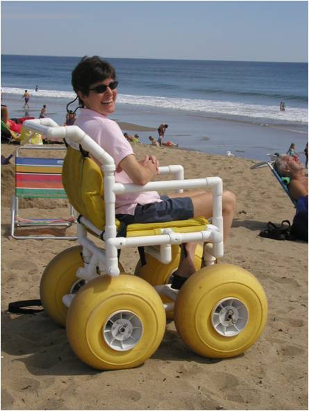 A smiling woman is sitting in a beach wheelchair made of PVC tubing, in front of the ocean. The chair has large yellow balloon tires and yellow cushions.