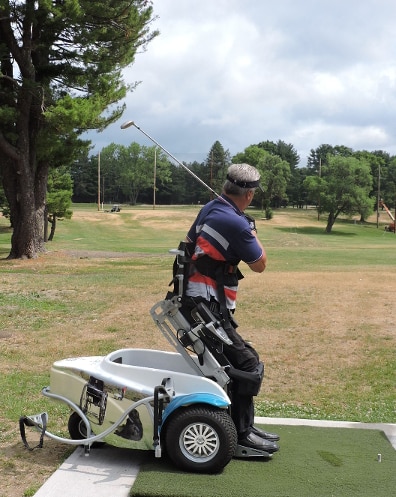 A man is using a ParaGolfer at the driving range. The ParaGolfer has a triangular base with two wheels in the front and one in the back. The seat is mounted on a hinged frame that can be raised. The man using the Paragolfer is strapped into ths seat with a harness, and is raised up to a standing position. His golf club is swung over his shoulder. he is looking down the driving range.